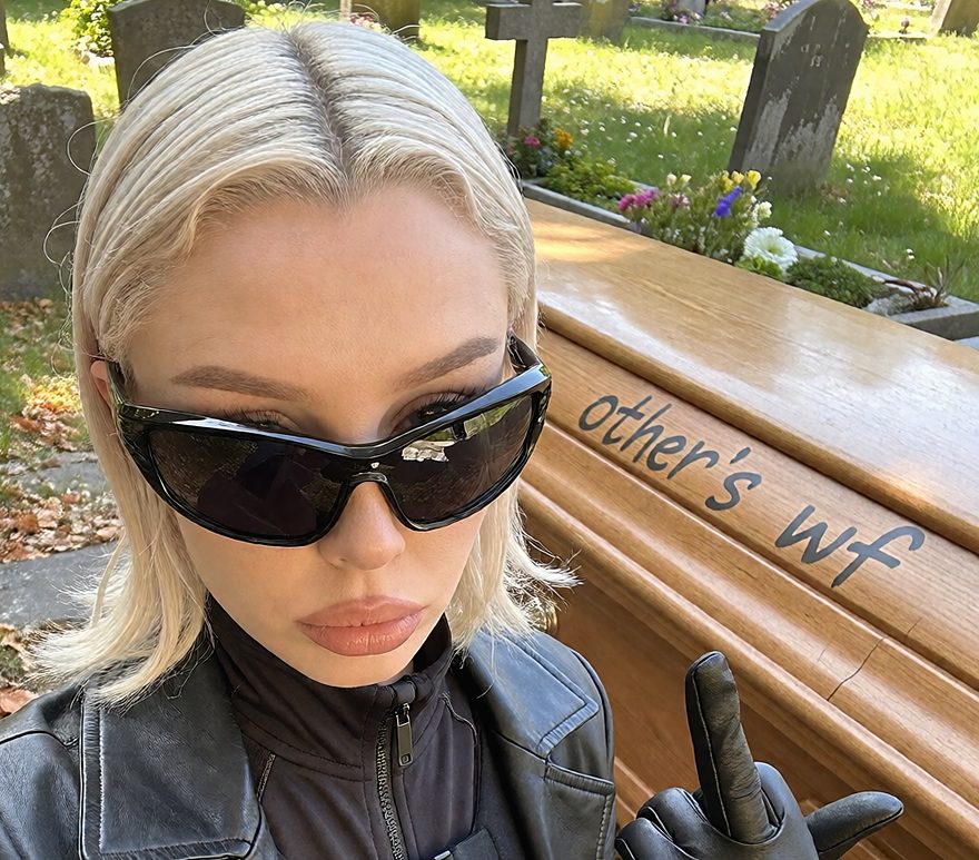 A blonde woman in sunglasses and a leather coat beside a coffin painted with the words other's wf.