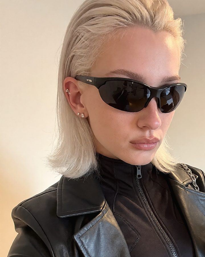 The same blonde woman in sunglasses and a leather coat, close up.